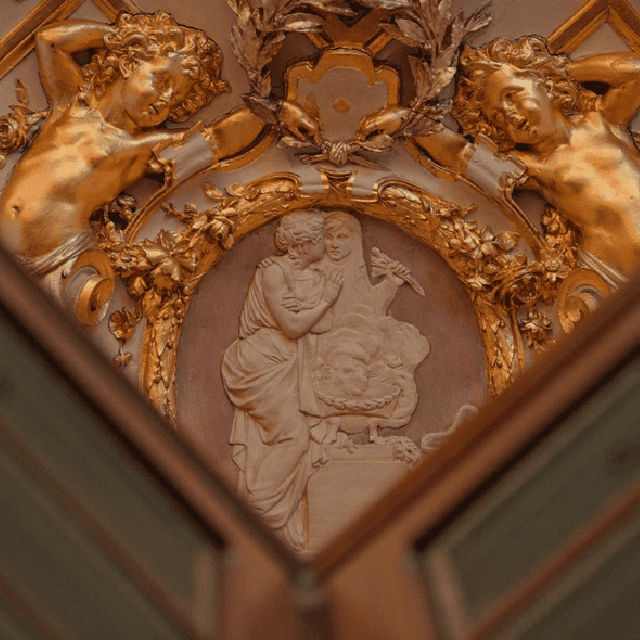 Alt text: Close-up of ornate ceiling decoration featuring cherubs and a central relief of a man and woman, surrounded by intricate floral motifs.