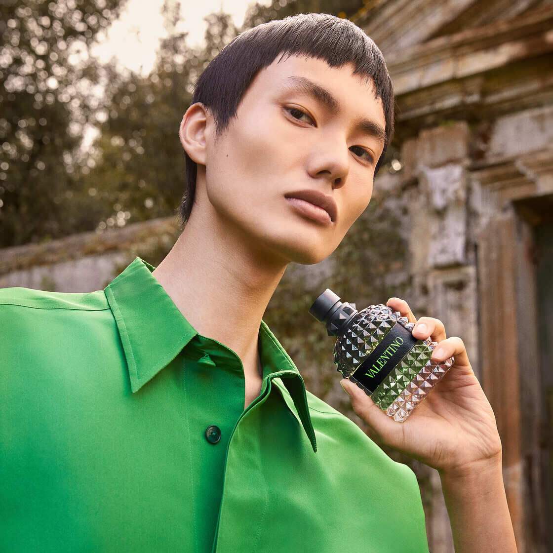 A model in a vibrant green shirt holds a Valentino perfume bottle, posing against a historic outdoor backdrop.