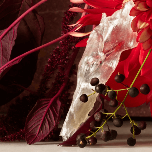 A close-up of a vibrant arrangement featuring deep red flowers, dark foliage, and an organic-looking clear sculpture, creating a striking contrast between natural and artistic elements.