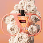 A bottle of Valentino perfume is surrounded by white roses, framed by a stylish pink "V" design, set against a soft gradient background.