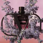 A stylish perfume bottle with a geometric design is framed by delicate purple flowers, set against a soft pink background. The label reads "Valentino."