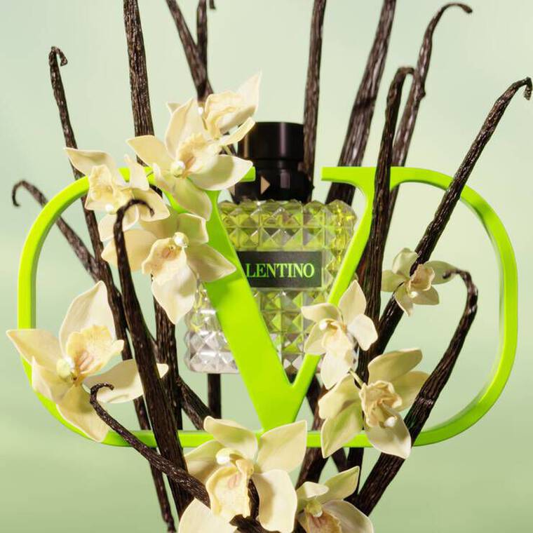 A bottle of Valentino perfume is surrounded by elegant white orchids and dark, twisted branches, set against a soft green background. The bottle is framed by a bold green logo resembling the letter "V."