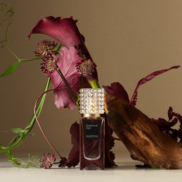 A Valentino perfume bottle placed beside deep red flowers and textured natural elements against a soft, light background.