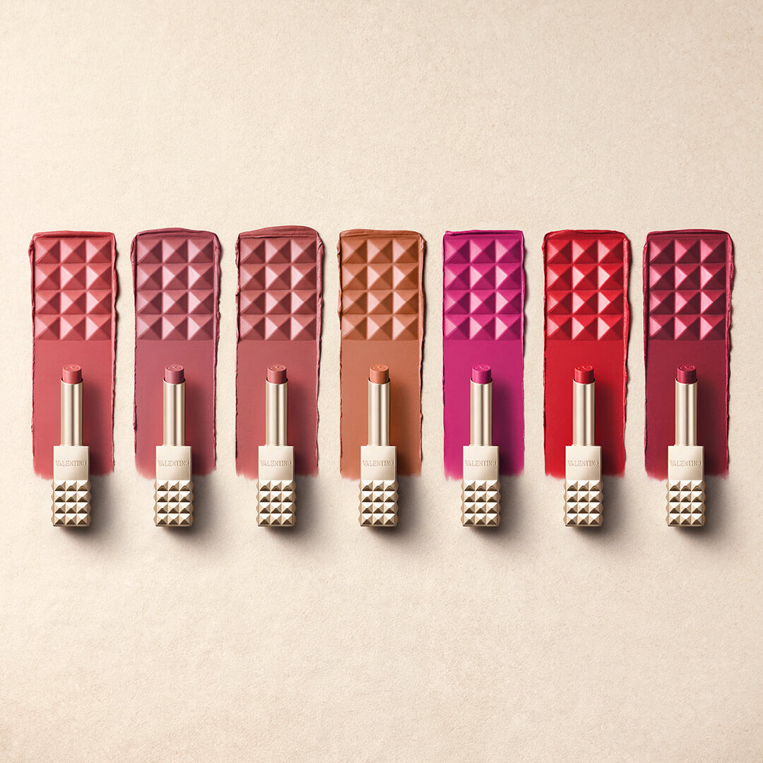 A collection of lipsticks in various shades displayed alongside matching swatches on a light background. The lipsticks are in sleek, textured packaging with pyramid-shaped accents.
