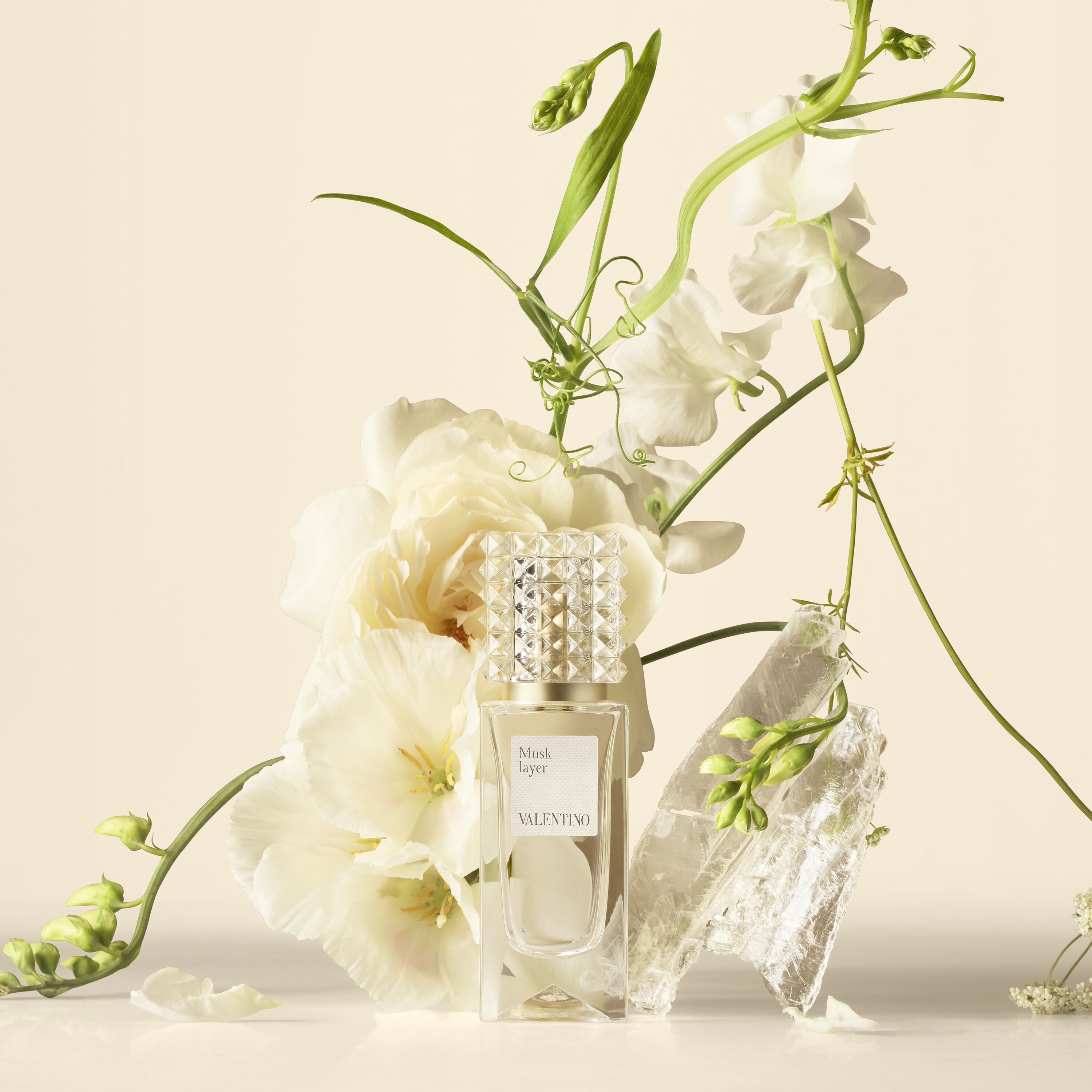 Un flacon de parfum Valentino accompagne de fleurs blanches et de cristaux translucides sur un fond neutre.