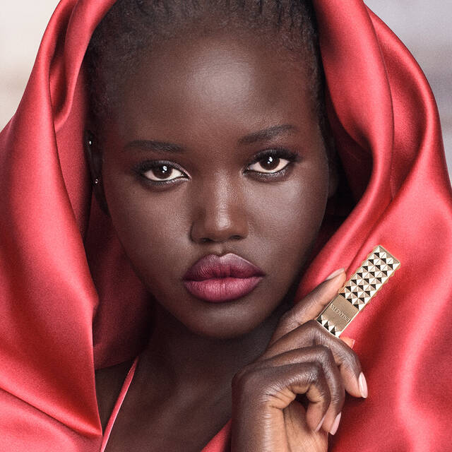 A model with deep brown skin and striking features poses confidently, wrapped in a vibrant red satin fabric. She holds a lipstick in her hand, showcasing a bold and glamorous look.