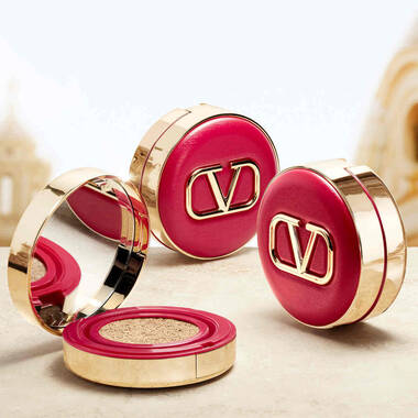 Three luxurious makeup compacts in vibrant red and gold, featuring the "DV" logo, with one compact open to reveal its mirrored interior and cushion foundation.