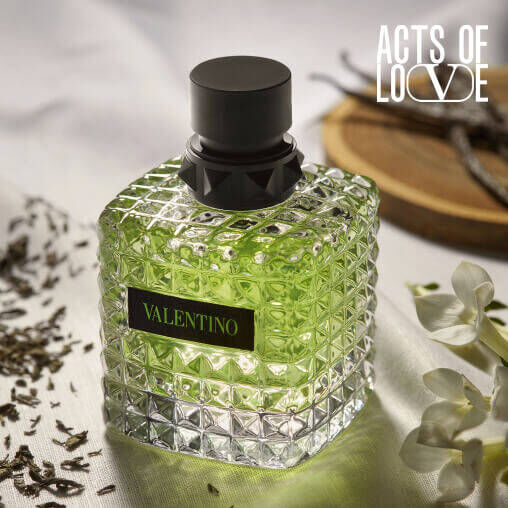 A green Valentino perfume bottle sits on a textured surface, surrounded by herbs and flowers, exuding elegance.