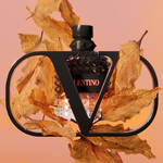 A bottle of Valentino perfume is surrounded by autumn leaves, set against a soft gradient background.