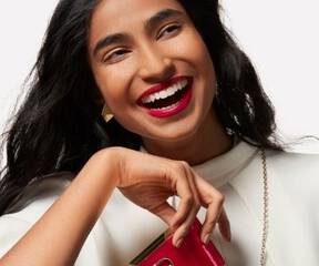A smiling woman with long, dark hair holding a small red purse, wearing bold red lipstick and elegant earrings, in front of a neutral background.