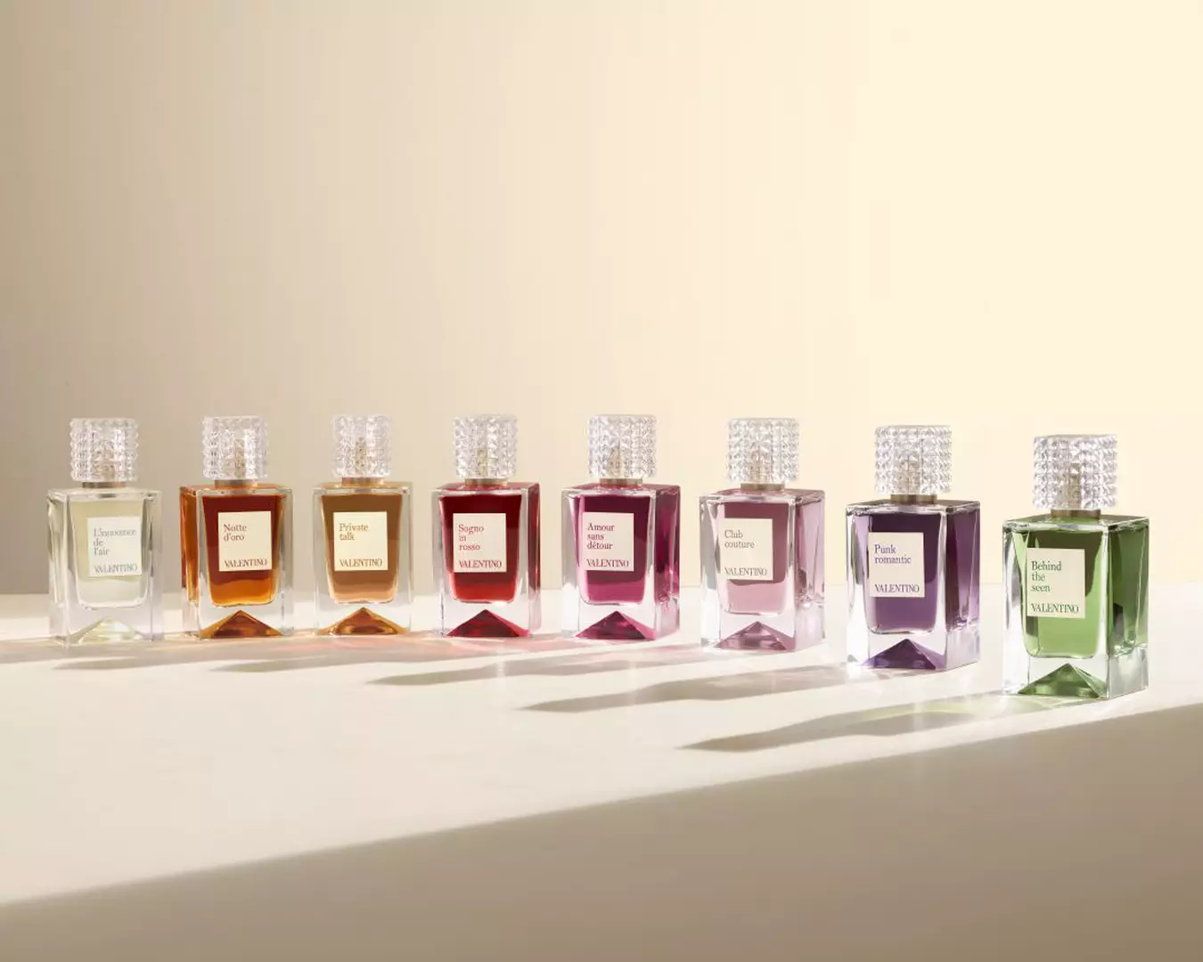 Eine Reihe von acht Valentino-Parfumflakons in verschiedenen Farben, jeweils mit einem strukturierten transparenten Verschluss, auf einer hellen Oberflache vor einem beigen Hintergrund angeordnet