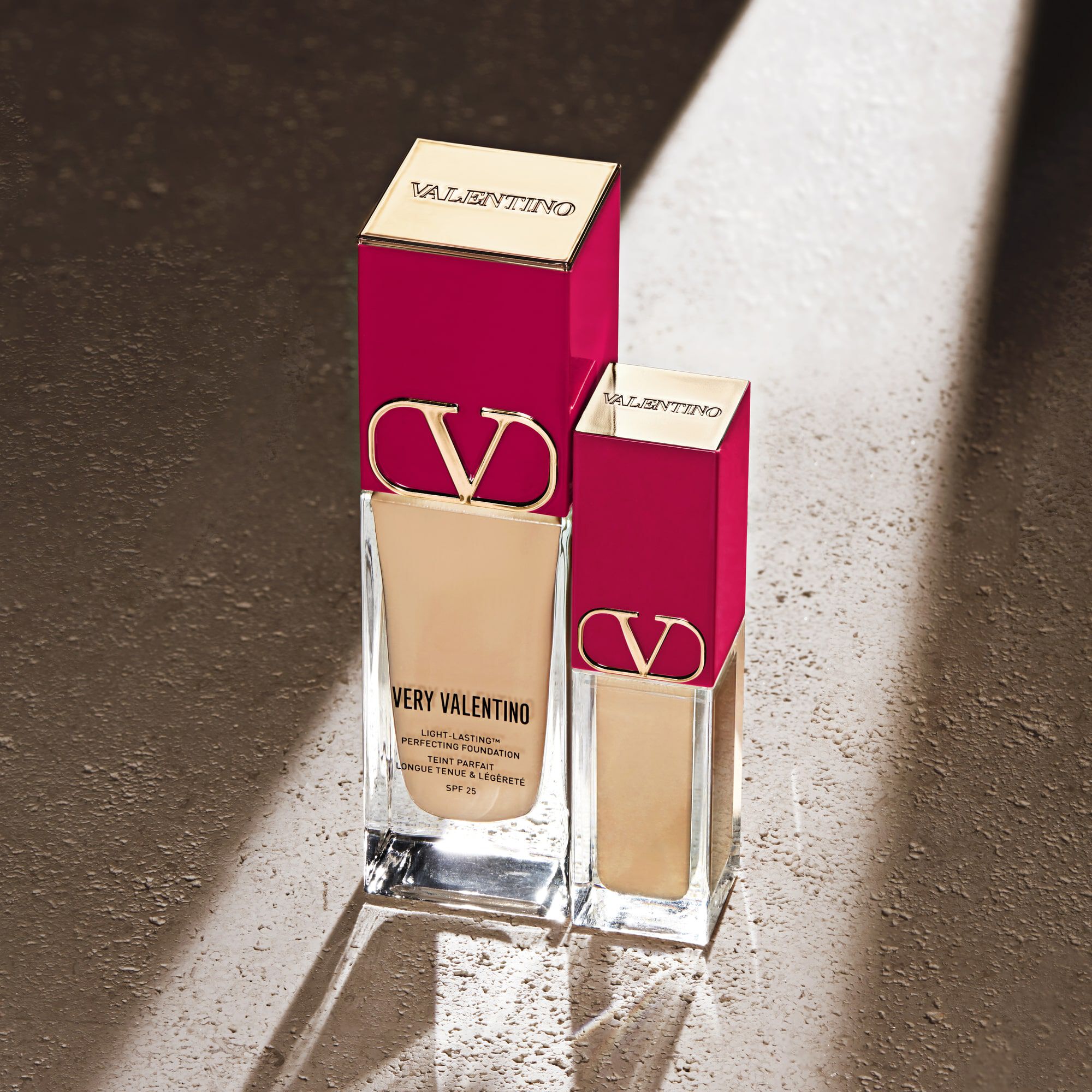 Two bottles of Very Valentino foundation in different sizes, elegantly displayed with a pink and gold design on a textured surface with soft lighting.