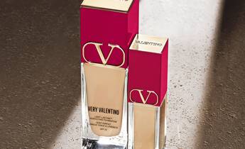 Two bottles of Very Valentino foundation in different sizes, elegantly displayed with a pink and gold design on a textured surface with soft lighting.