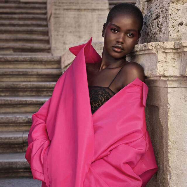 A model poses elegantly in a vibrant pink oversized jacket, set against a stone stairway background.