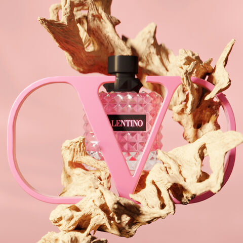 A stylish pink perfume bottle enclosed in a large, sculptural letter "V" against a soft pink background, surrounded by organic, textured wood elements.