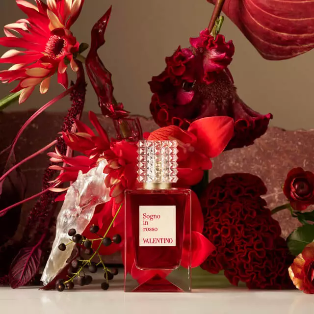 A bottle of Valentino perfume labeled "Sogno in Rosso" surrounded by vibrant red flowers and rich maroon textures, creating an elegant and luxurious aesthetic.