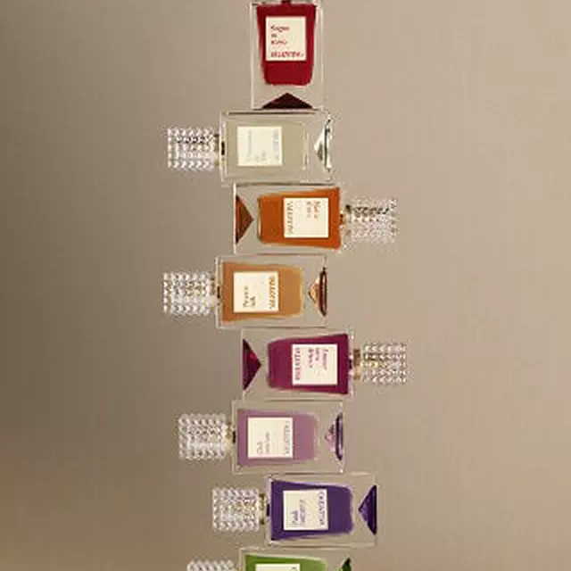A row of eight Valentino perfume bottles in different colors, each with a textured transparent cap, arranged on a light surface against a beige background.