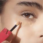 A close-up of a woman applying concealer under her eye with a wand applicator. Her skin is glowing, and she has natural eyelashes.