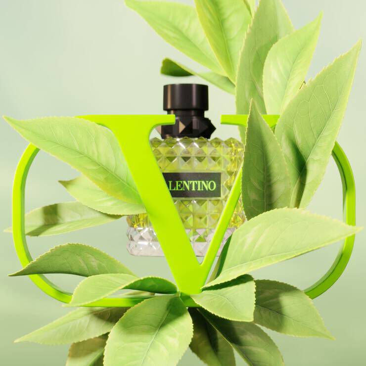 A diamond-shaped perfume bottle with a black cap, labeled "Valentino," surrounded by vibrant green leaves and framed by a bold green "V." The background is a soft green hue.