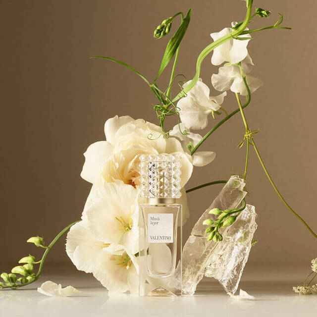 A Valentino perfume bottle arranged with white flowers and translucent crystals on a neutral background.