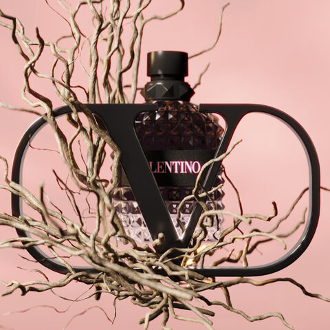 A stylized perfume bottle with a diamond-patterned design is framed by a black, abstract structure, surrounded by twisted, bare branches against a soft pink background. The label reads "Valentino."
