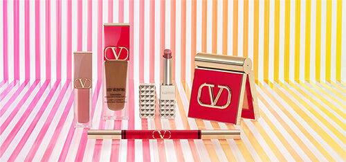 A collection of luxury makeup products arranged on a striped gradient background featuring colors from pink to yellow. The products include lipsticks, a foundation, and a compact, all showcasing elegant packaging.