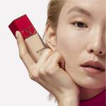 A close-up of a model holding a bottle of foundation with a striking pink and gold design, showcasing a natural makeup look.