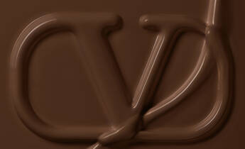 A close-up of a smooth, melted chocolate surface with a stylized logo imprinted in it. The logo features the letters "CD" intricately designed in a flowing, chocolate-like texture.