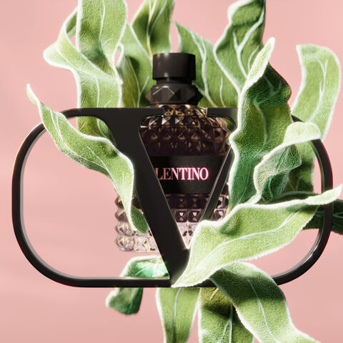 A stylish perfume bottle with a black diamond-patterned design is surrounded by vibrant green leaves, all set against a soft pink background. The bottle features the brand name "Valentino" prominently displayed.