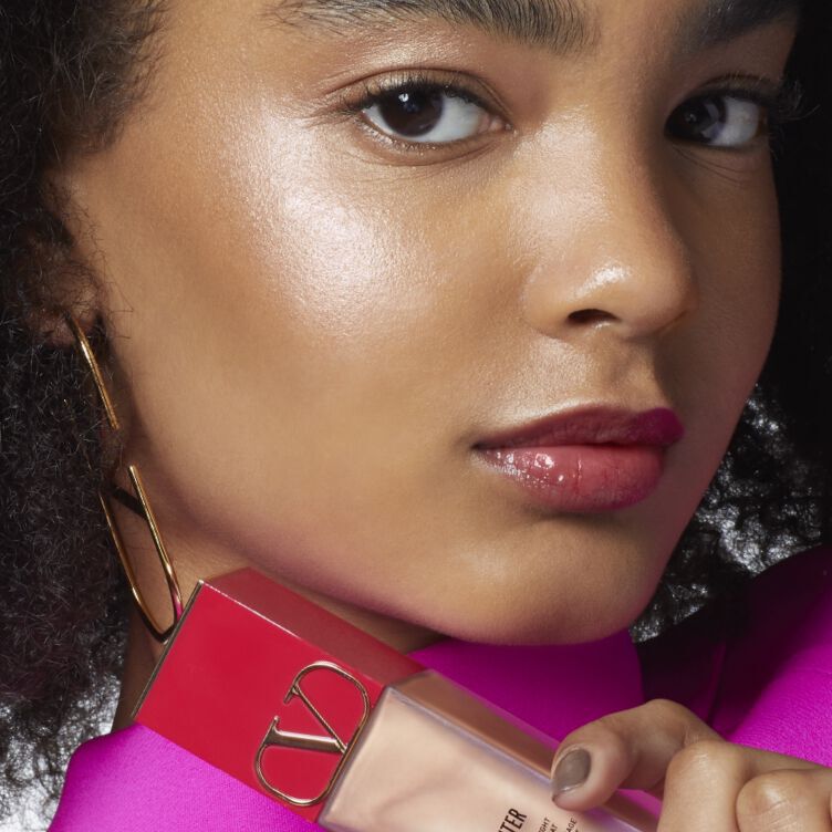 A close-up of a young woman with curly black hair, wearing gold hoop earrings and a bright pink top. She holds a bottle of foundation, looking confidently at the camera with a subtle glossy lip.
