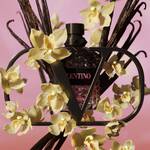 A decorative perfume bottle is surrounded by delicate yellow orchids and dark branches, set against a soft pink background. The bottle has a geometric design and features the brand name "Valentino."
